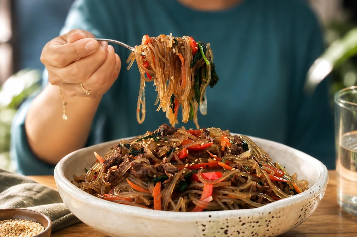 A person using a fork to lift a bite of Japchae from a large bowl. A person using a fork to lift a bite of Japchae from a large bowl.