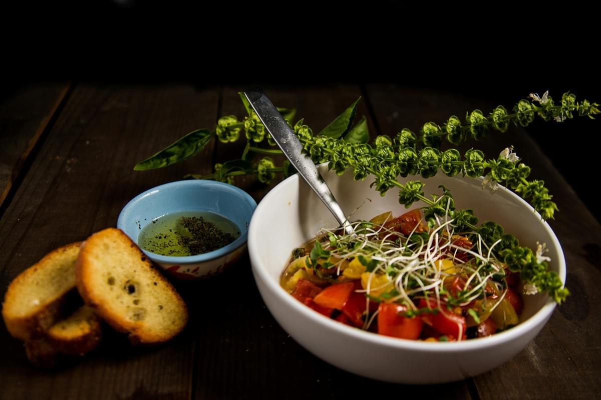 A white bowl filled with diced red and yellow tomatoes, microgreens, and fresh basil sprigs sits on a dark wooden table alongside a spoon. Beside the bowl, three slices of toasted crostini rest next to a small blue dish containing olive oil and black pepper. A white bowl filled with diced red and yellow tomatoes, microgreens, and fresh basil sprigs sits on a dark wooden table alongside a spoon. Beside the bowl, three slices of toasted crostini rest next to a small blue dish containing olive oil and black pepper.