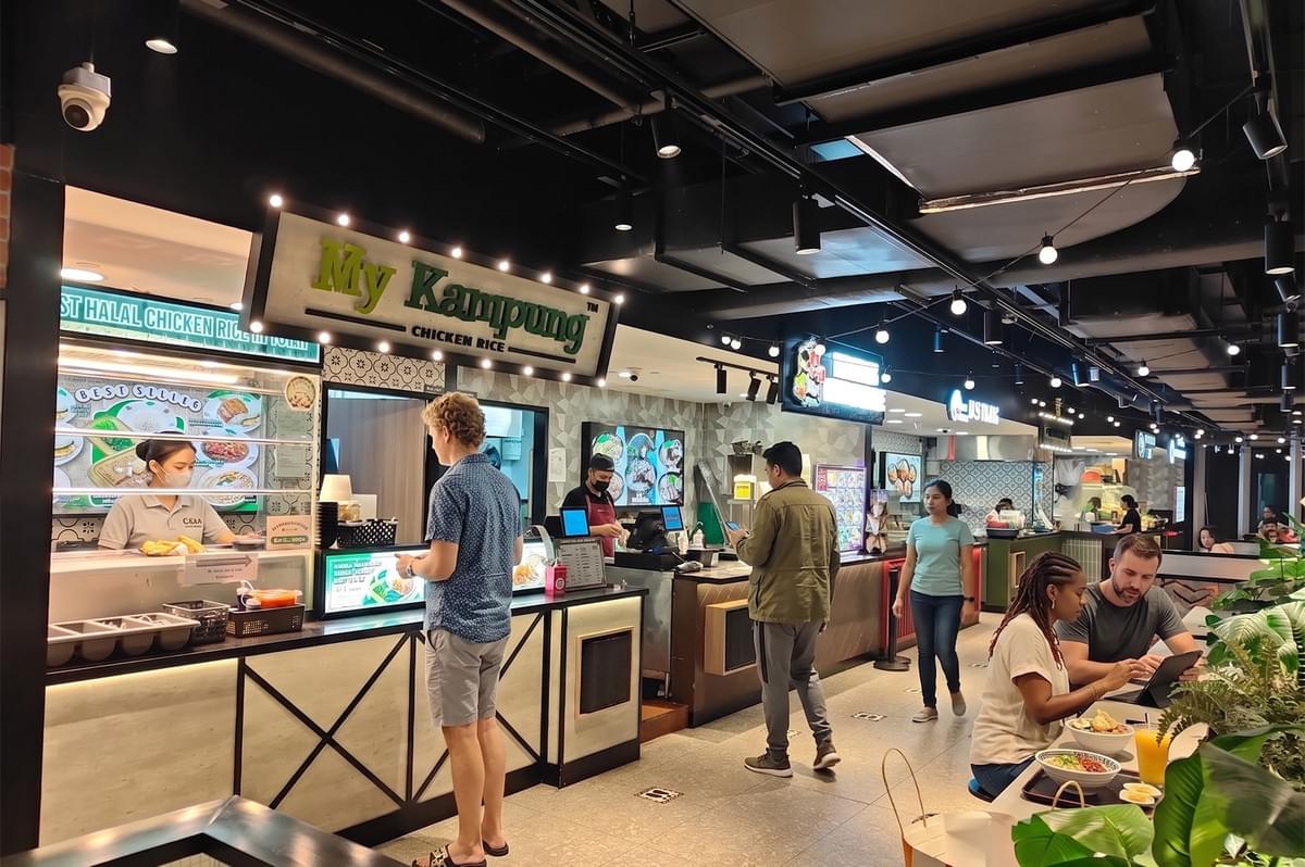 A modern food court interior featuring a My Kampung Chicken Rice stall with customers ordering and dining in a clean, well-lit environment. A modern food court interior featuring a My Kampung Chicken Rice stall with customers ordering and dining in a clean, well-lit environment.