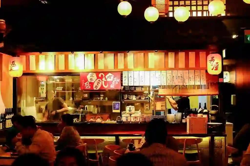 A warm, dimly lit Japanese izakaya features a busy open kitchen where chefs prepare food behind a counter lined with lanterns and menu signs. Customers sit at the bar and surrounding wooden tables, enjoying their meal under the soft glow of hanging spherical lights. A warm, dimly lit Japanese izakaya features a busy open kitchen where chefs prepare food behind a counter lined with lanterns and menu signs. Customers sit at the bar and surrounding wooden tables, enjoying their meal under the soft glow of hanging spherical lights.