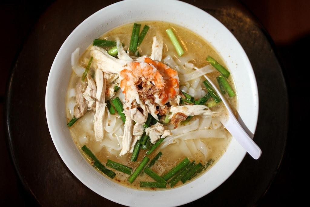 A white bowl filled with noodle soup features flat rice noodles, shredded chicken, and a couple of cooked shrimp topped with fried shallots. The soup is garnished with chopped green onions and served with a white plastic spoon, set against a dark background. A white bowl filled with noodle soup features flat rice noodles, shredded chicken, and a couple of cooked shrimp topped with fried shallots. The soup is garnished with chopped green onions and served with a white plastic spoon, set against a dark background.