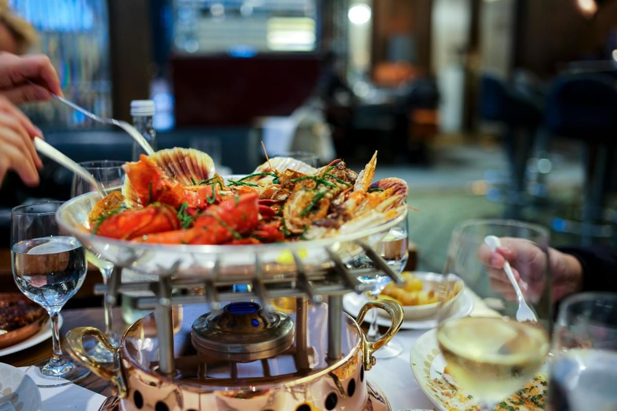 A platter of cooked seafood, including lobster, scallops, and prawns, sits on a raised burner in a restaurant. Diners reach in with utensils around glasses of wine and water on the table. A platter of cooked seafood, including lobster, scallops, and prawns, sits on a raised burner in a restaurant. Diners reach in with utensils around glasses of wine and water on the table.