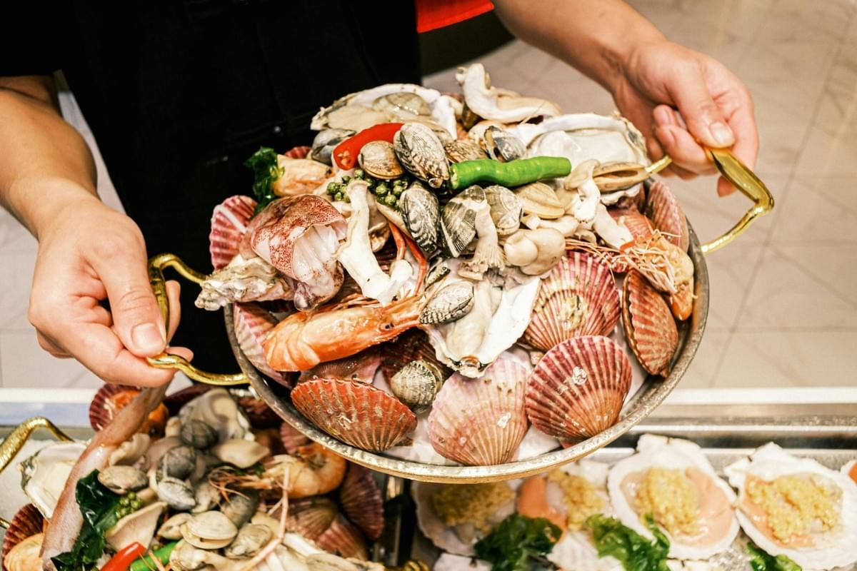 A person holds a large, round metal platter overflowing with an assortment of fresh seafood, including scallops, clams, shrimp, squid, and chili peppers. Additional prepared seafood dishes sit displayed on the counter directly below. A person holds a large, round metal platter overflowing with an assortment of fresh seafood, including scallops, clams, shrimp, squid, and chili peppers. Additional prepared seafood dishes sit displayed on the counter directly below.