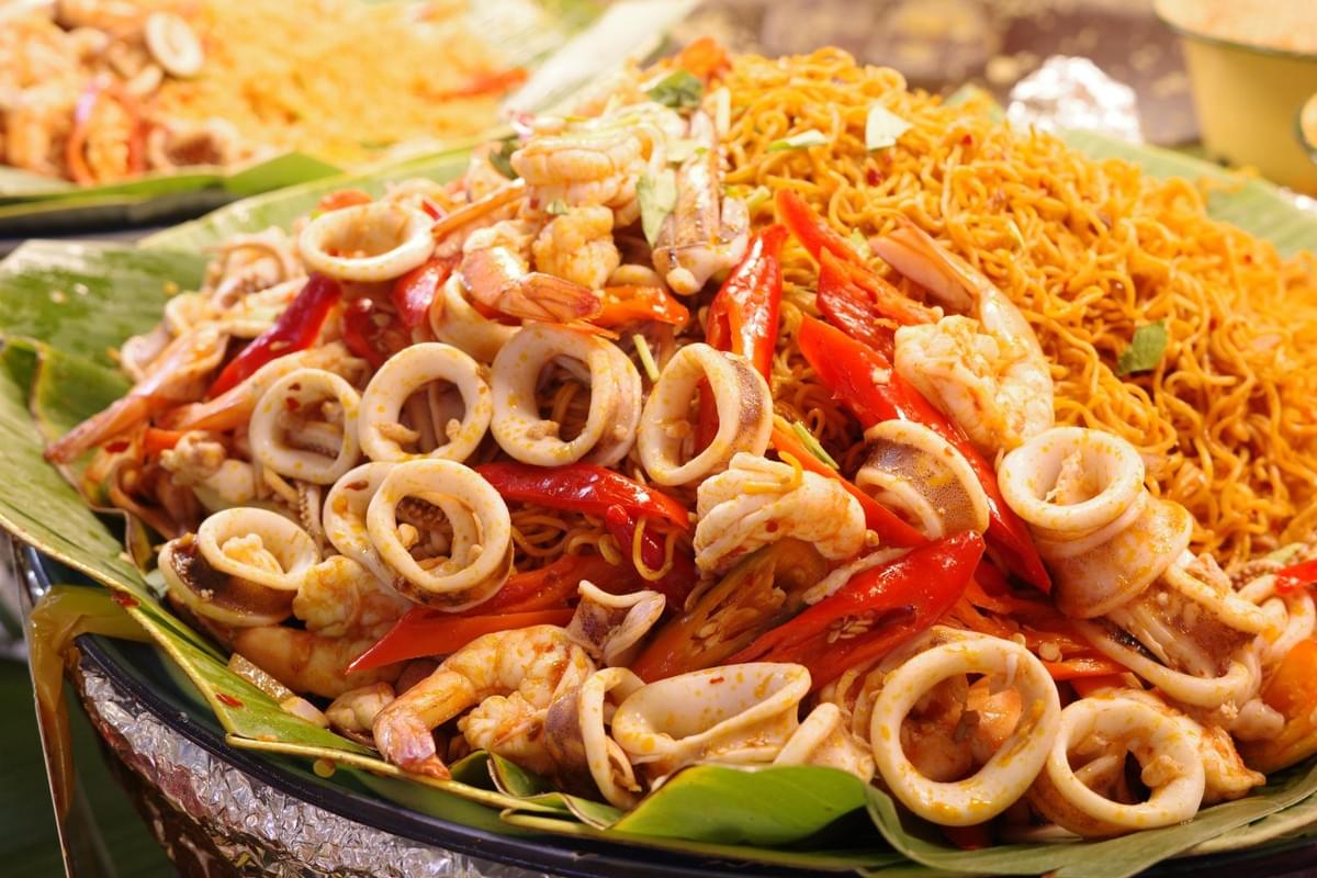 A platter served on banana leaves is piled high with spicy fried stir-fried noodles topped with squid rings, shrimp, and red chili peppers. The colorful dish showcases fresh seafood tossed together in a flavorful street food style presentation. A platter served on banana leaves is piled high with spicy fried stir-fried noodles topped with squid rings, shrimp, and red chili peppers. The colorful dish showcases fresh seafood tossed together in a flavorful street food style presentation.