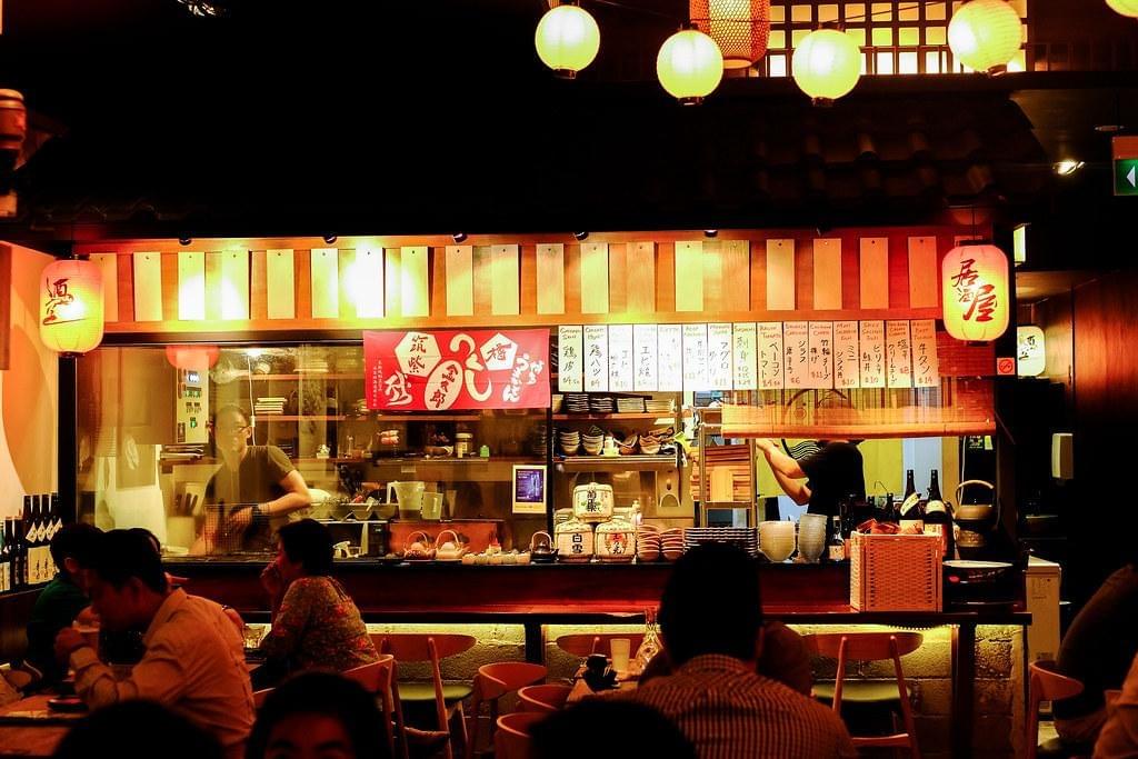 A warm, dimly lit Japanese izakaya features a busy open kitchen where chefs prepare food behind a counter lined with lanterns and menu signs. Customers sit at the bar and surrounding wooden tables, enjoying their meal under the soft glow of hanging spherical lights. A warm, dimly lit Japanese izakaya features a busy open kitchen where chefs prepare food behind a counter lined with lanterns and menu signs. Customers sit at the bar and surrounding wooden tables, enjoying their meal under the soft glow of hanging spherical lights.