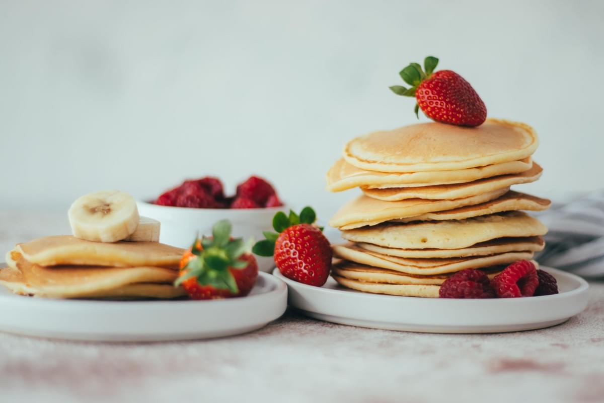 A tall stack of fluffy, golden-brown pancakes sits on a white plate, topped with a whole fresh strawberry and accompanied by loose raspberries at the base. Next to it, a smaller stack of pancakes is topped with sliced bananas and served alongside fresh strawberries and a small bowl of berries against a bright, neutral background. A tall stack of fluffy, golden-brown pancakes sits on a white plate, topped with a whole fresh strawberry and accompanied by loose raspberries at the base. Next to it, a smaller stack of pancakes is topped with sliced bananas and served alongside fresh strawberries and a small bowl of berries against a bright, neutral background.