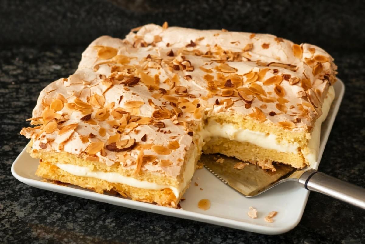 Rectangular Kvæfjordkake meringue cake on a platter with a missing slice being lifted with a spatula. Rectangular Kvæfjordkake meringue cake on a platter with a missing slice being lifted with a spatula.