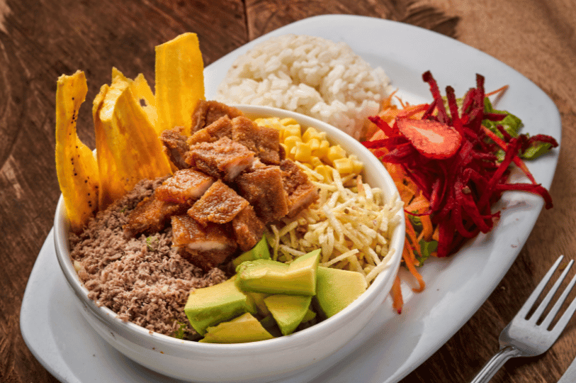 A modern presentation of Bandeja Paisa ingredients served "bowl-style," featuring chopped chicharrón, ground meat, avocado, corn, and thin plantain chips, accompanied by rice and a beet salad. A modern presentation of Bandeja Paisa ingredients served "bowl-style," featuring chopped chicharrón, ground meat, avocado, corn, and thin plantain chips, accompanied by rice and a beet salad.