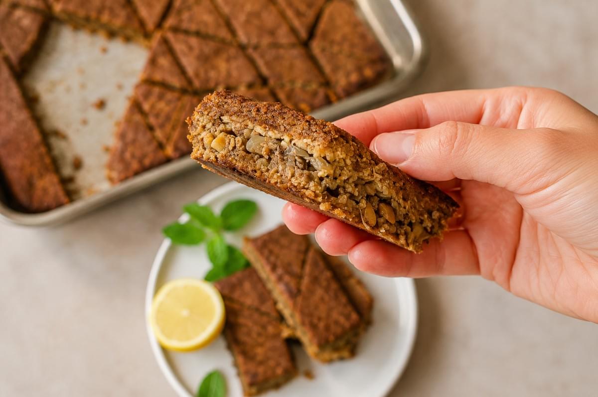A hand holding a piece of baked kibbeh bil sanieh, showing its spiced meat and pine nut filling layer, with a slice of lemon nearby. A hand holding a piece of baked kibbeh bil sanieh, showing its spiced meat and pine nut filling layer, with a slice of lemon nearby.