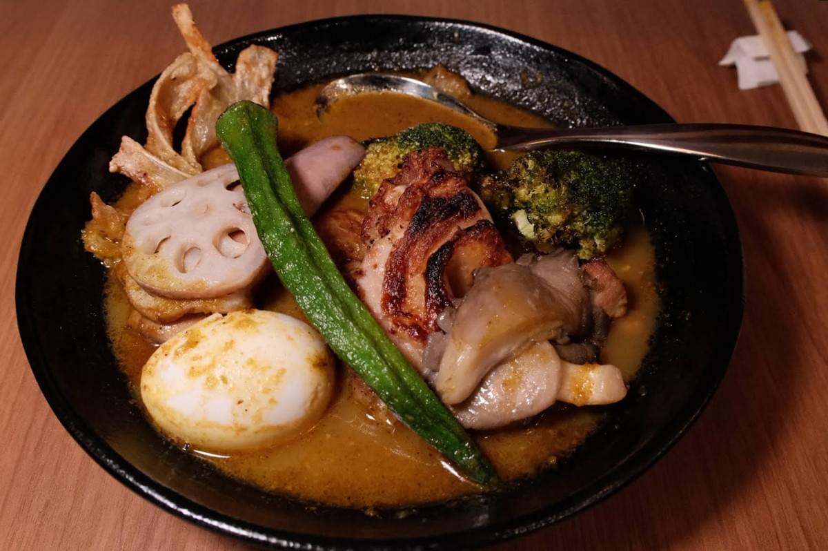 An overhead shot shows a dark ceramic bowl filled with a rich Hokkaido-style soup curry served with various colorful toppings. The dish includes grilled chicken, a whole soft-boiled egg, fried lotus root slices, a long green okra pod, charred broccoli florets, mushrooms, and crispy chips submerged in a savory broth, set on a warm wooden dining table alongside chopsticks. An overhead shot shows a dark ceramic bowl filled with a rich Hokkaido-style soup curry served with various colorful toppings. The dish includes grilled chicken, a whole soft-boiled egg, fried lotus root slices, a long green okra pod, charred broccoli florets, mushrooms, and crispy chips submerged in a savory broth, set on a warm wooden dining table alongside chopsticks.