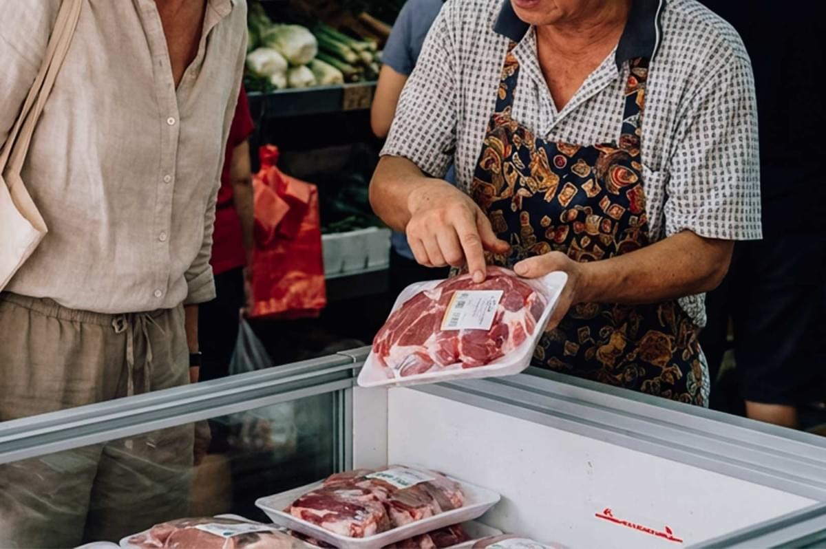 At a market stall, a vendor wearing a patterned apron and short-sleeved shirt holds up a plastic-wrapped tray of raw meat, pointing toward its label. Standing across from the vendor, a customer wearing a beige linen shirt and trousers looks down at the meat product above an open refrigerated display case filled with additional packaged cuts. At a market stall, a vendor wearing a patterned apron and short-sleeved shirt holds up a plastic-wrapped tray of raw meat, pointing toward its label. Standing across from the vendor, a customer wearing a beige linen shirt and trousers looks down at the meat product above an open refrigerated display case filled with additional packaged cuts.