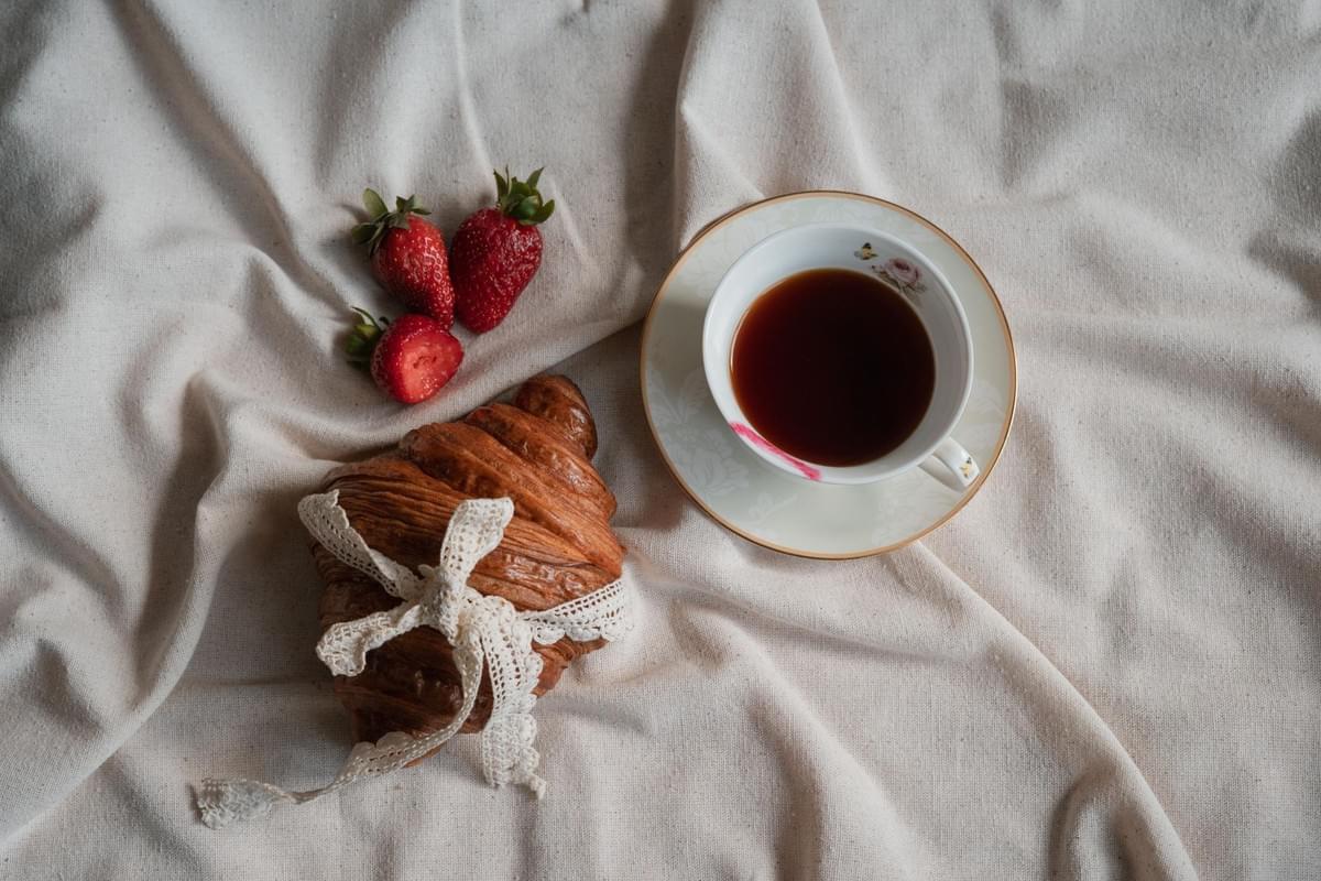 An overhead shot shows a cozy breakfast arranged on a rumpled, off-white fabric sheet. Next to a white teacup filled with dark tea are three fresh strawberries and a golden-brown croissant elegantly wrapped in a white lace bow. An overhead shot shows a cozy breakfast arranged on a rumpled, off-white fabric sheet. Next to a white teacup filled with dark tea are three fresh strawberries and a golden-brown croissant elegantly wrapped in a white lace bow.