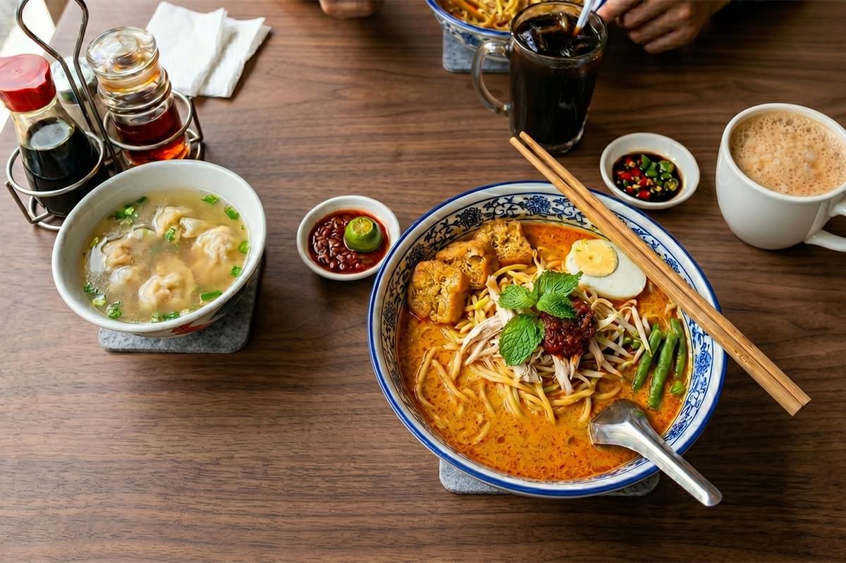 A bowl of curry laksa noodles served with a side of wonton soup, chili paste, and teh tarik coffee on a wooden table. A bowl of curry laksa noodles served with a side of wonton soup, chili paste, and teh tarik coffee on a wooden table.