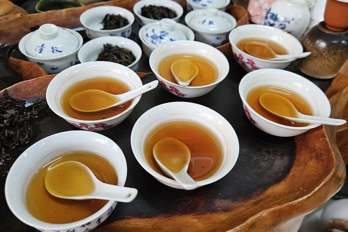A wooden tray holds multiple white ceramic bowls filled with amber tea, each containing a small white tasting spoon. Behind them, small covered gaiwans and cups filled with loose tea leaves are arranged for a tea cupping session. A wooden tray holds multiple white ceramic bowls filled with amber tea, each containing a small white tasting spoon. Behind them, small covered gaiwans and cups filled with loose tea leaves are arranged for a tea cupping session.