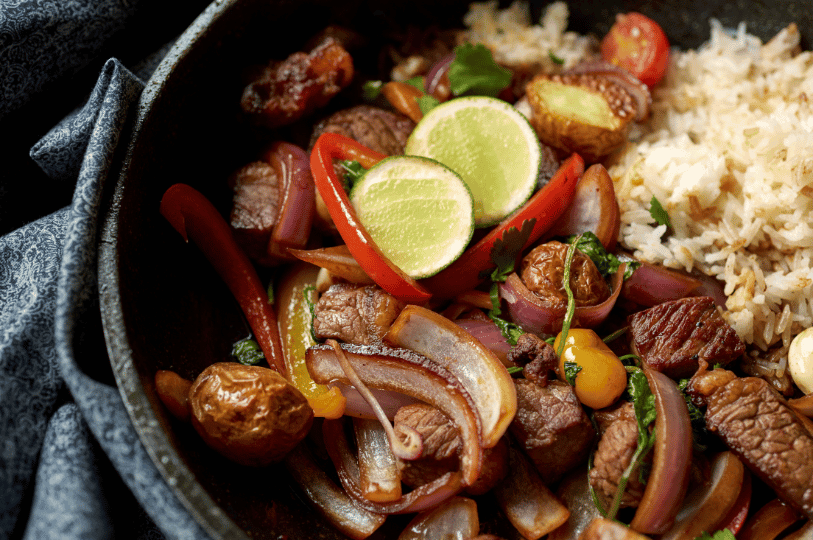 A close-up, top-down view of Lomo Saltado served in a dark cast-iron pan. The dish features seared beef chunks, red onions, and bell peppers, garnished with fresh lime slices and served alongside fluffy white rice. A close-up, top-down view of Lomo Saltado served in a dark cast-iron pan. The dish features seared beef chunks, red onions, and bell peppers, garnished with fresh lime slices and served alongside fluffy white rice.
