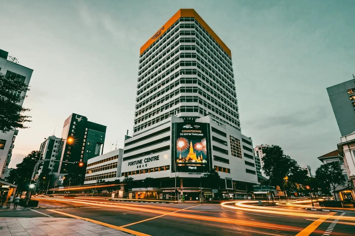 A high-angle, long-exposure photograph captures the tall Fortune Centre building with its distinctive white façade and yellow roof during twilight. Bright light trails from passing traffic streak across the street intersection in the foreground. A high-angle, long-exposure photograph captures the tall Fortune Centre building with its distinctive white façade and yellow roof during twilight. Bright light trails from passing traffic streak across the street intersection in the foreground.
