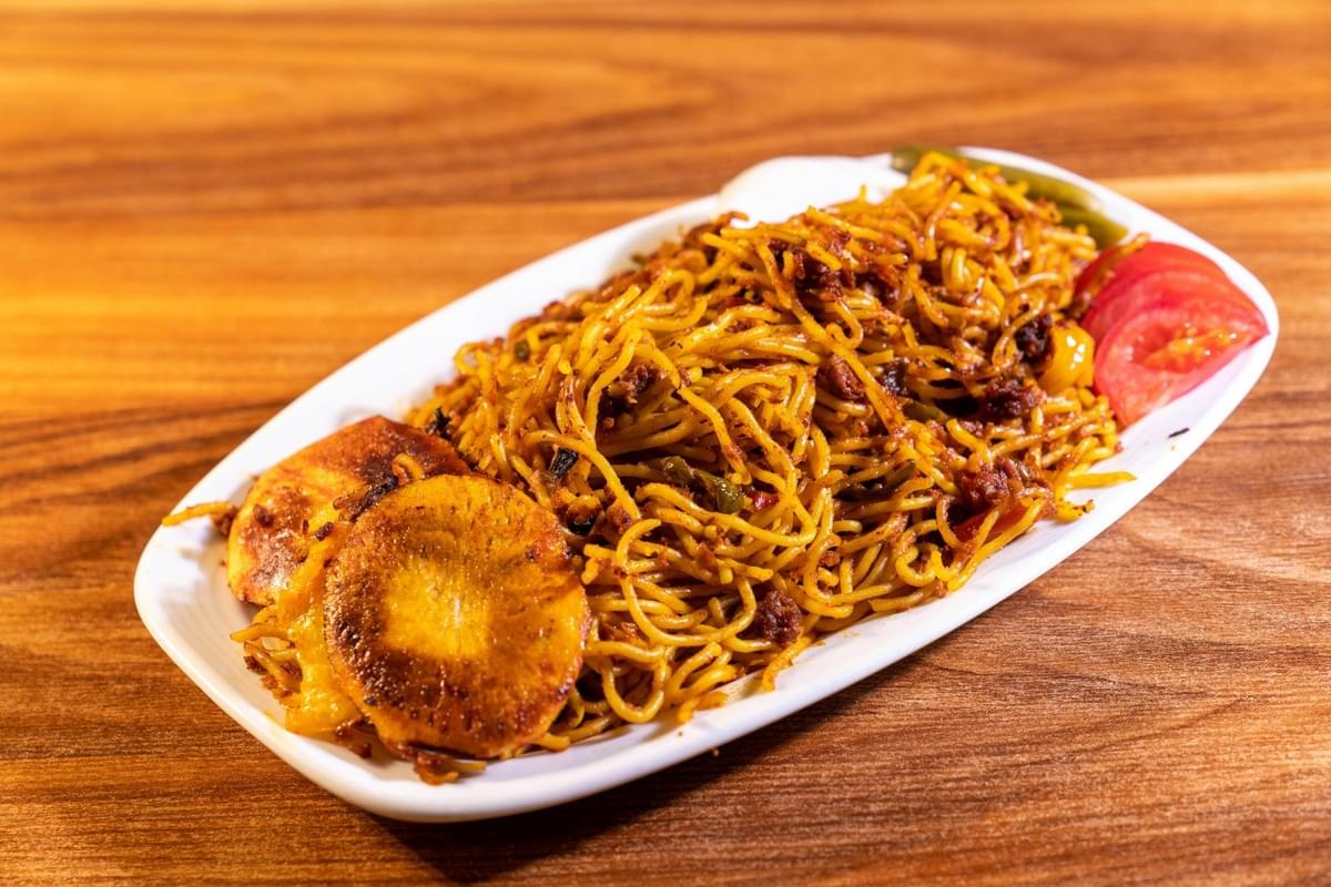A platter of seasoned, golden-brown noodles is served on a white dish alongside crispy, fried rounds of potato and sliced tomatoes. The entire meal sits on a warm-toned wooden tabletop background. A platter of seasoned, golden-brown noodles is served on a white dish alongside crispy, fried rounds of potato and sliced tomatoes. The entire meal sits on a warm-toned wooden tabletop background.
