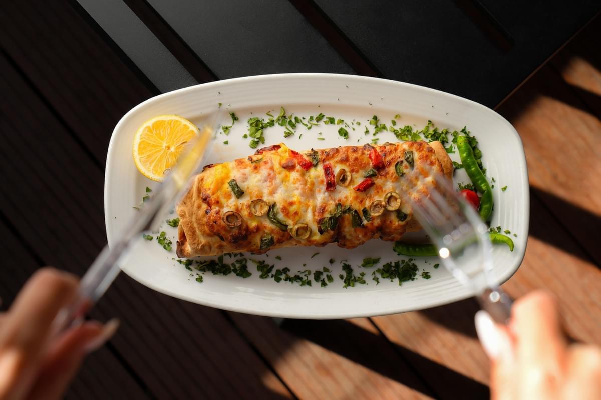 An overhead shot of the image shows a baked burrito-style dish covered in melted cheese, sliced olives, and peppers, served on an oval plate garnished with herbs and a lemon wheel. In the foreground, a person's hands hold a clear knife and fork, slightly blurred, as they prepare to eat the meal on an outdoor wooden deck. An overhead shot of the image shows a baked burrito-style dish covered in melted cheese, sliced olives, and peppers, served on an oval plate garnished with herbs and a lemon wheel. In the foreground, a person's hands hold a clear knife and fork, slightly blurred, as they prepare to eat the meal on an outdoor wooden deck.