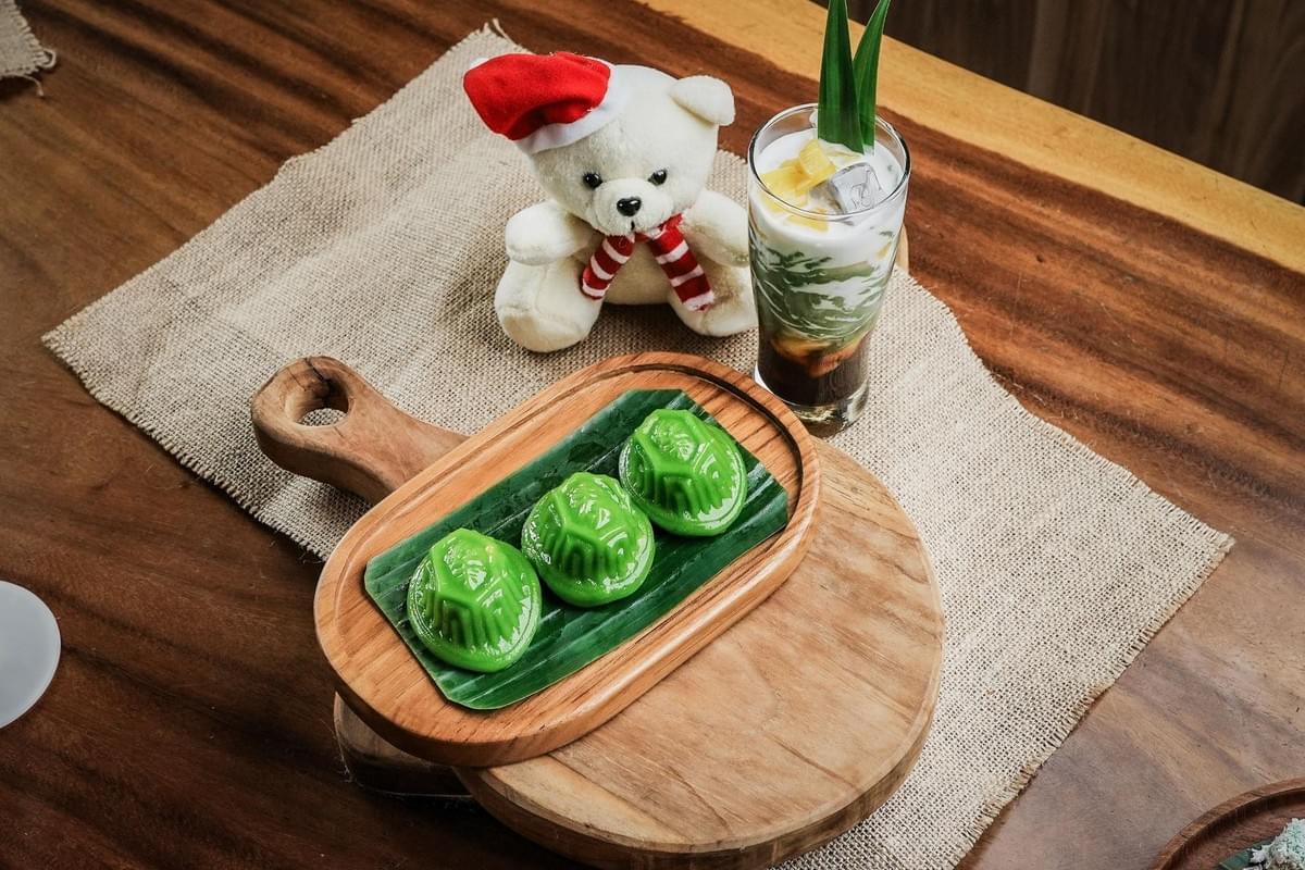 Three molded green kueh cakes are served on a banana leaf atop a wooden board alongside a tall glass of layered iced cendol. In the background, a small plush teddy bear wearing a Santa hat and scarf sits on a burlap placemat. Three molded green kueh cakes are served on a banana leaf atop a wooden board alongside a tall glass of layered iced cendol. In the background, a small plush teddy bear wearing a Santa hat and scarf sits on a burlap placemat.
