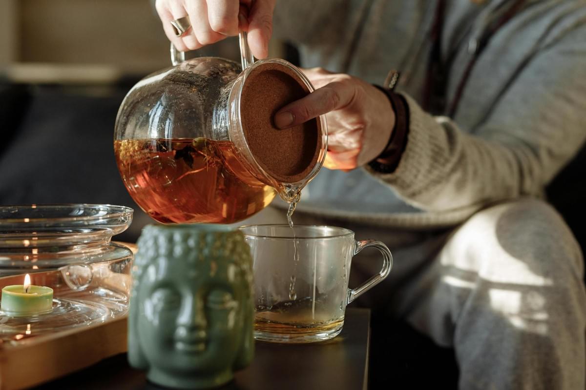 Bathed in warm sunlight, a person pours amber herbal tea from a round glass teapot with a cork lid into a matching clear cup. In the foreground, a serene green Buddha head statue rests beside a lit candle warmer, creating a peaceful and spiritual atmosphere for the tea session. Bathed in warm sunlight, a person pours amber herbal tea from a round glass teapot with a cork lid into a matching clear cup. In the foreground, a serene green Buddha head statue rests beside a lit candle warmer, creating a peaceful and spiritual atmosphere for the tea session.