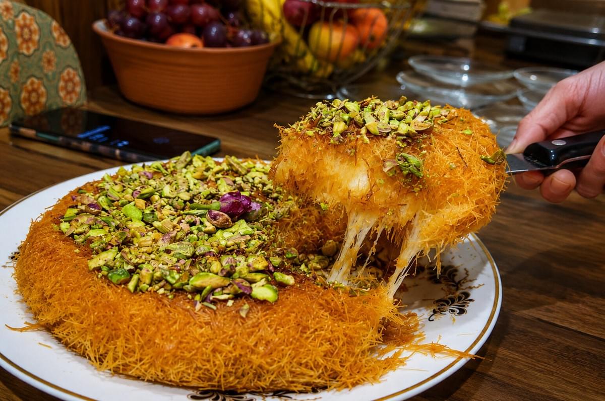 A hand uses a server to lift a slice of golden, crispy kunafa pastry from a large platter, creating a thick stretch of melted cheese, with chopped pistachios generously scattered over the top. A hand uses a server to lift a slice of golden, crispy kunafa pastry from a large platter, creating a thick stretch of melted cheese, with chopped pistachios generously scattered over the top.