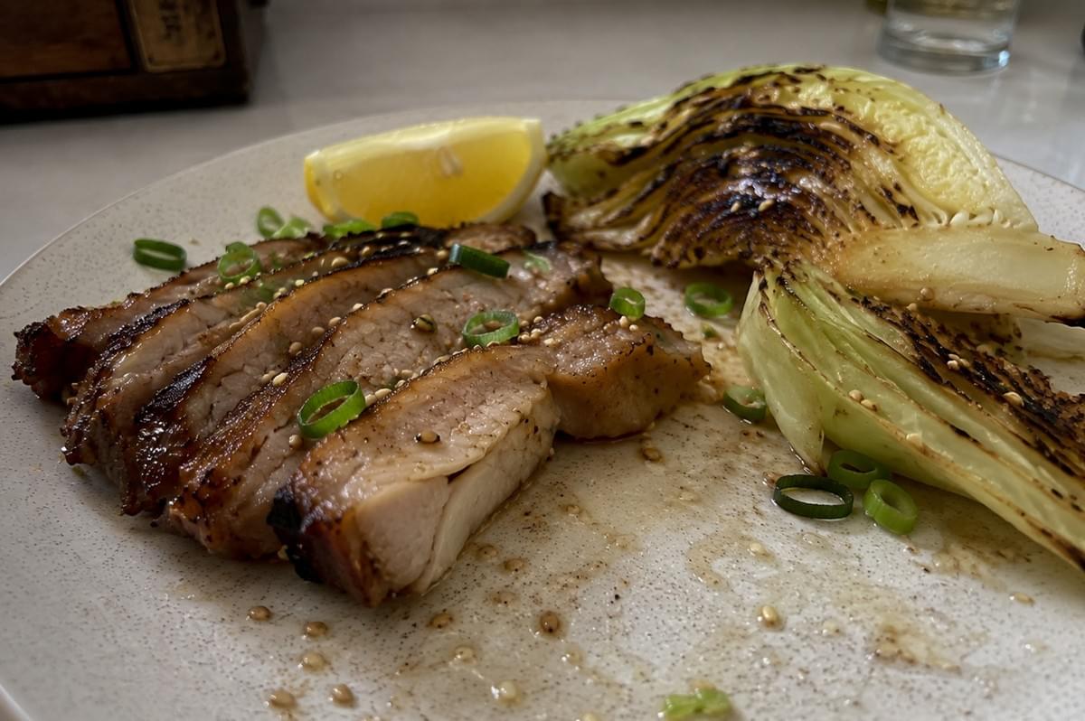 A close-up, eye-level shot of a partially plated dish on a speckled ceramic plate, featuring a fan of sliced, grilled pork belly on the left and charred cabbage wedges on the right. The pork belly slices display a beautifully seared, dark brown edge and are garnished with scattered sesame seeds and bright green scallion rings, with a single lemon wedge resting behind them. Juices and sesame seeds are speckled across the empty space of the plate in the foreground, capturing an authentic, documentary-style look at the meal preparation. A close-up, eye-level shot of a partially plated dish on a speckled ceramic plate, featuring a fan of sliced, grilled pork belly on the left and charred cabbage wedges on the right. The pork belly slices display a beautifully seared, dark brown edge and are garnished with scattered sesame seeds and bright green scallion rings, with a single lemon wedge resting behind them. Juices and sesame seeds are speckled across the empty space of the plate in the foreground, capturing an authentic, documentary-style look at the meal preparation.