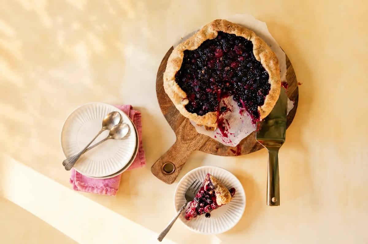 An overhead shot of a rustic mixed berry galette on a wooden cutting board, with one slice cut out and served on a small white plate nearby. Two empty white plates with spoons resting on a pink napkin are placed to the left under soft, warm sunlight. An overhead shot of a rustic mixed berry galette on a wooden cutting board, with one slice cut out and served on a small white plate nearby. Two empty white plates with spoons resting on a pink napkin are placed to the left under soft, warm sunlight.