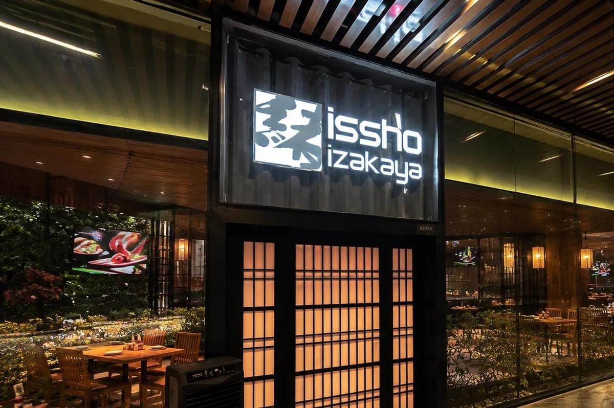 An illuminated "issho izakaya" sign hangs above a traditional shoji-style entrance door of a Japanese restaurant. Large glass windows flank the entrance, revealing warmly lit wooden tables and chairs set for dining inside. An illuminated "issho izakaya" sign hangs above a traditional shoji-style entrance door of a Japanese restaurant. Large glass windows flank the entrance, revealing warmly lit wooden tables and chairs set for dining inside.