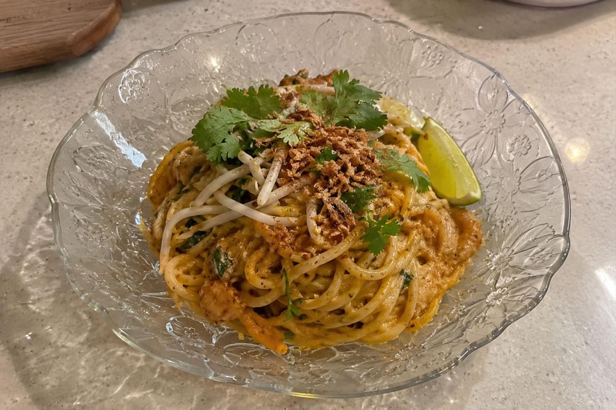 A high-angle shot captures a bowl of creamy, orange-hued laksa carbonara pasta served in a decorative clear glass bowl on a countertop. The spaghetti is coated in rich sauce and garnished with fresh cilantro leaves, crunchy bean sprouts, crispy fried shallots, and a lime wedge on the rim. A high-angle shot captures a bowl of creamy, orange-hued laksa carbonara pasta served in a decorative clear glass bowl on a countertop. The spaghetti is coated in rich sauce and garnished with fresh cilantro leaves, crunchy bean sprouts, crispy fried shallots, and a lime wedge on the rim.