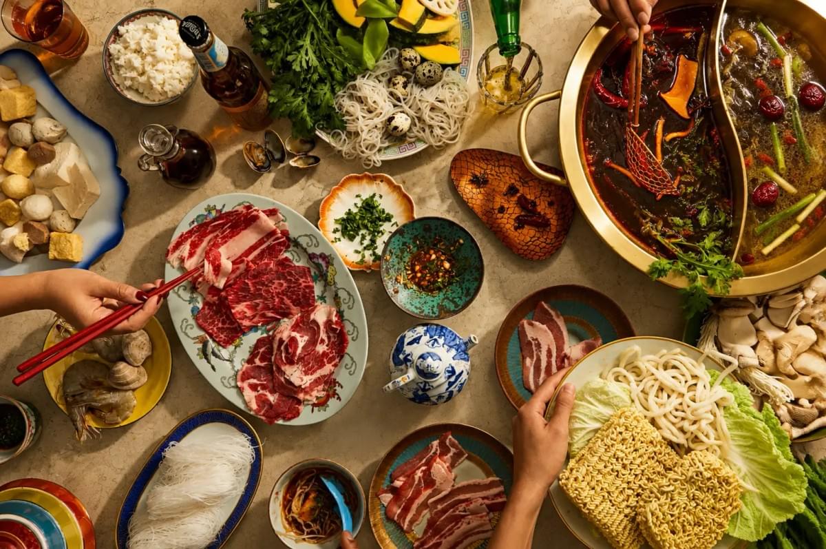 Overhead view of people using chopsticks to cook sliced meat in a two-flavor hot pot surrounded by side ingredients. Overhead view of people using chopsticks to cook sliced meat in a two-flavor hot pot surrounded by side ingredients.