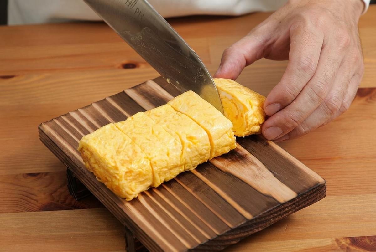 Chef slicing rolled Japanese omelet (tamagoyaki) on a wooden board with a large knife. Chef slicing rolled Japanese omelet (tamagoyaki) on a wooden board with a large knife.