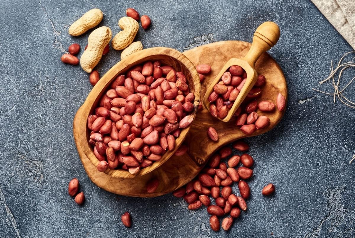 Flat lay of raw red-skin peanuts in a wooden bowl with a scoop on a dark surface. Flat lay of raw red-skin peanuts in a wooden bowl with a scoop on a dark surface.