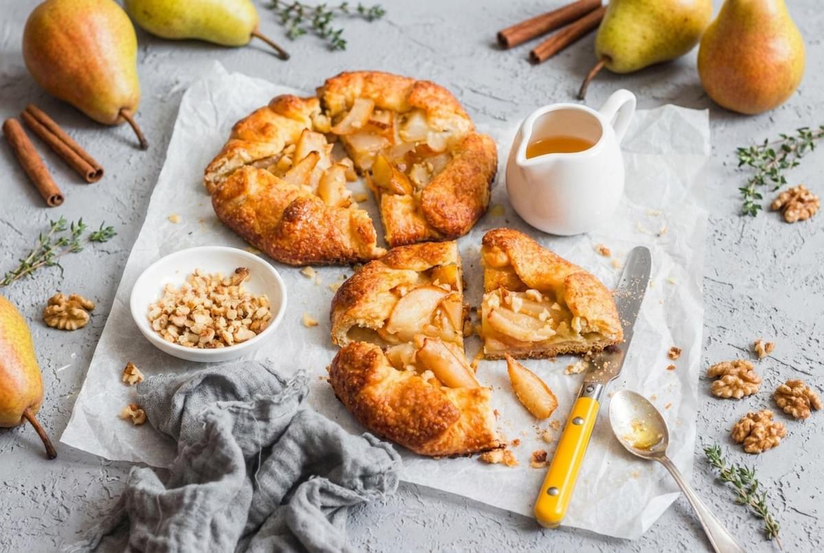 Slices of golden-brown pear galette on parchment paper, served with chopped walnuts, a small honey pitcher, and a yellow knife. Slices of golden-brown pear galette on parchment paper, served with chopped walnuts, a small honey pitcher, and a yellow knife.
