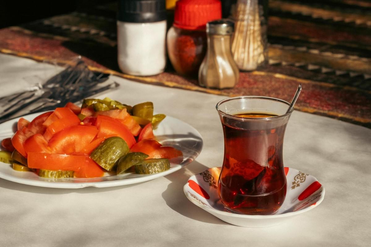 Bathed in bright sunlight, a traditional tulip-shaped glass of dark tea sits on a saucer beside a plate of sliced tomatoes and pickles. The outdoor dining setting includes cutlery and condiment shakers resting on a white tablecloth with a patterned runner. Bathed in bright sunlight, a traditional tulip-shaped glass of dark tea sits on a saucer beside a plate of sliced tomatoes and pickles. The outdoor dining setting includes cutlery and condiment shakers resting on a white tablecloth with a patterned runner.