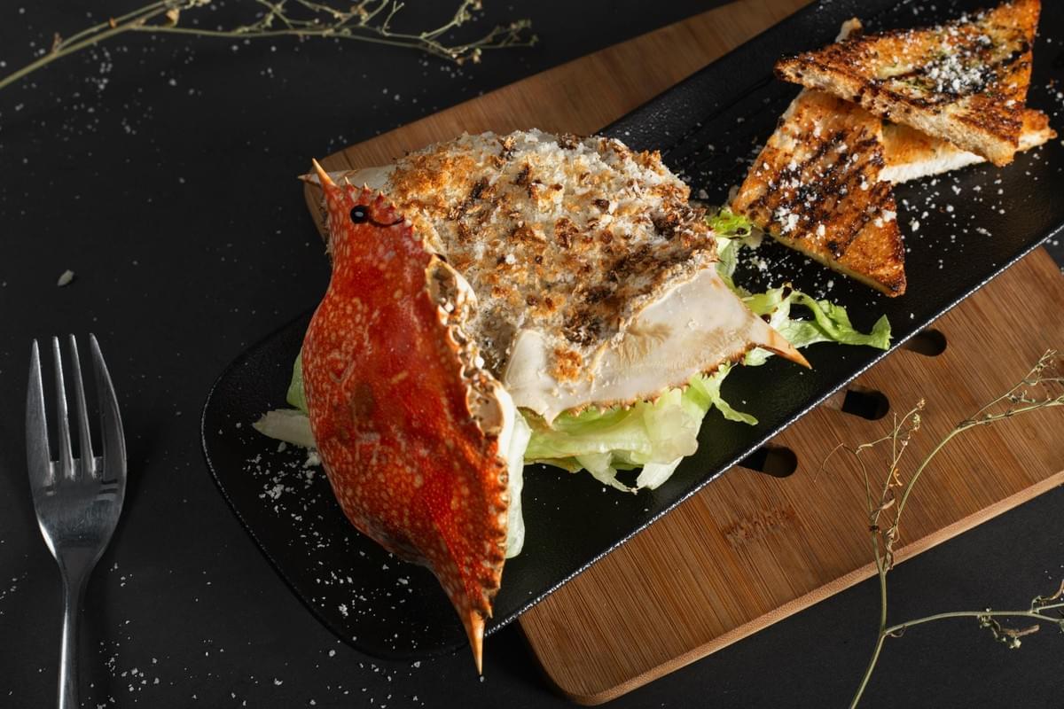 A baked stuffed crab dish served over lettuce leaves sits on a long black serving tray alongside triangular toasted bread points. The dish is presented on a wooden board against a dark background with a metal fork resting to the side. A baked stuffed crab dish served over lettuce leaves sits on a long black serving tray alongside triangular toasted bread points. The dish is presented on a wooden board against a dark background with a metal fork resting to the side.