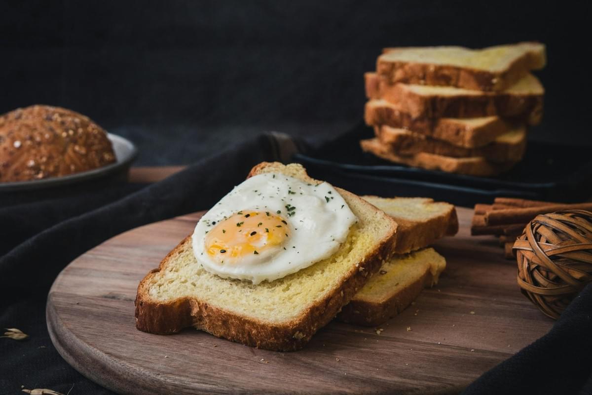 A sunny-side-up fried egg garnished with herbs sits atop a slice of toasted white bread on a round wooden cutting board. In the dark-toned background, a stack of toasted bread, a whole grain roll, and cinnamon sticks complete the cozy breakfast scene. A sunny-side-up fried egg garnished with herbs sits atop a slice of toasted white bread on a round wooden cutting board. In the dark-toned background, a stack of toasted bread, a whole grain roll, and cinnamon sticks complete the cozy breakfast scene.