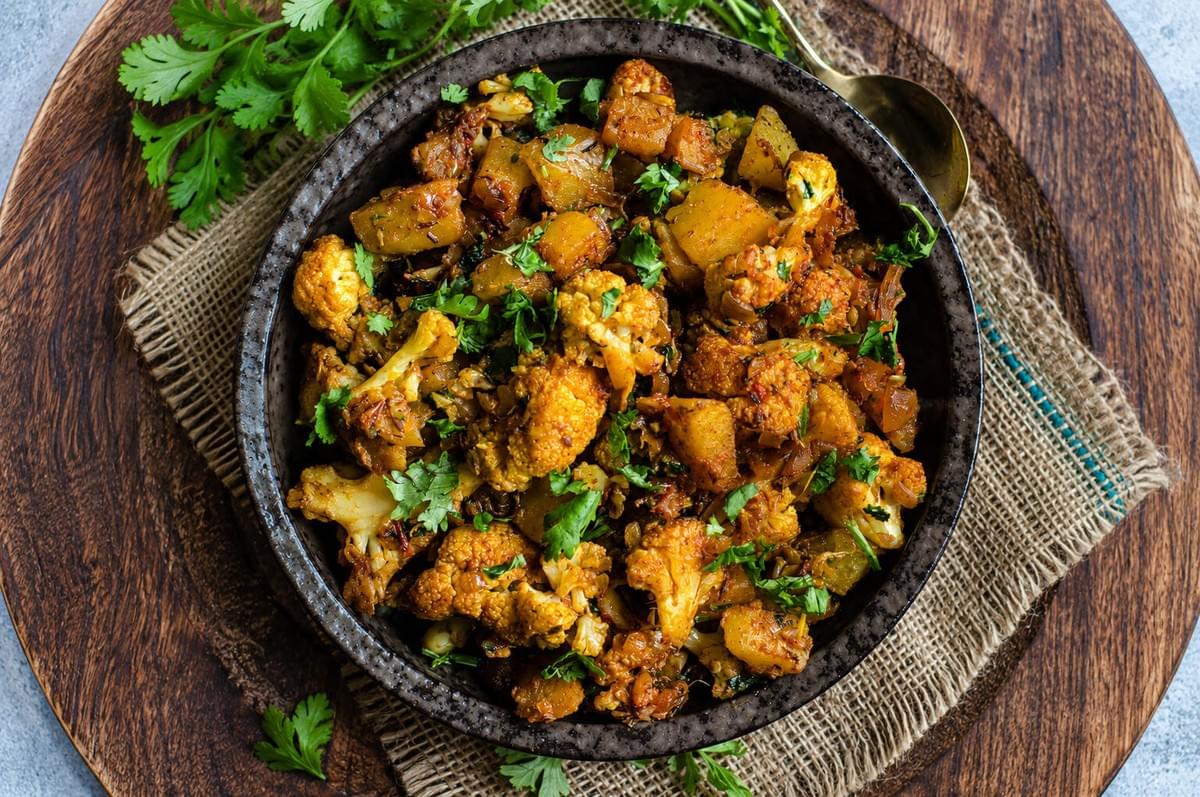 Overhead view of Aloo Gobi topped with chopped cilantro in a dark stoneware bowl set on a wooden charger. Overhead view of Aloo Gobi topped with chopped cilantro in a dark stoneware bowl set on a wooden charger.