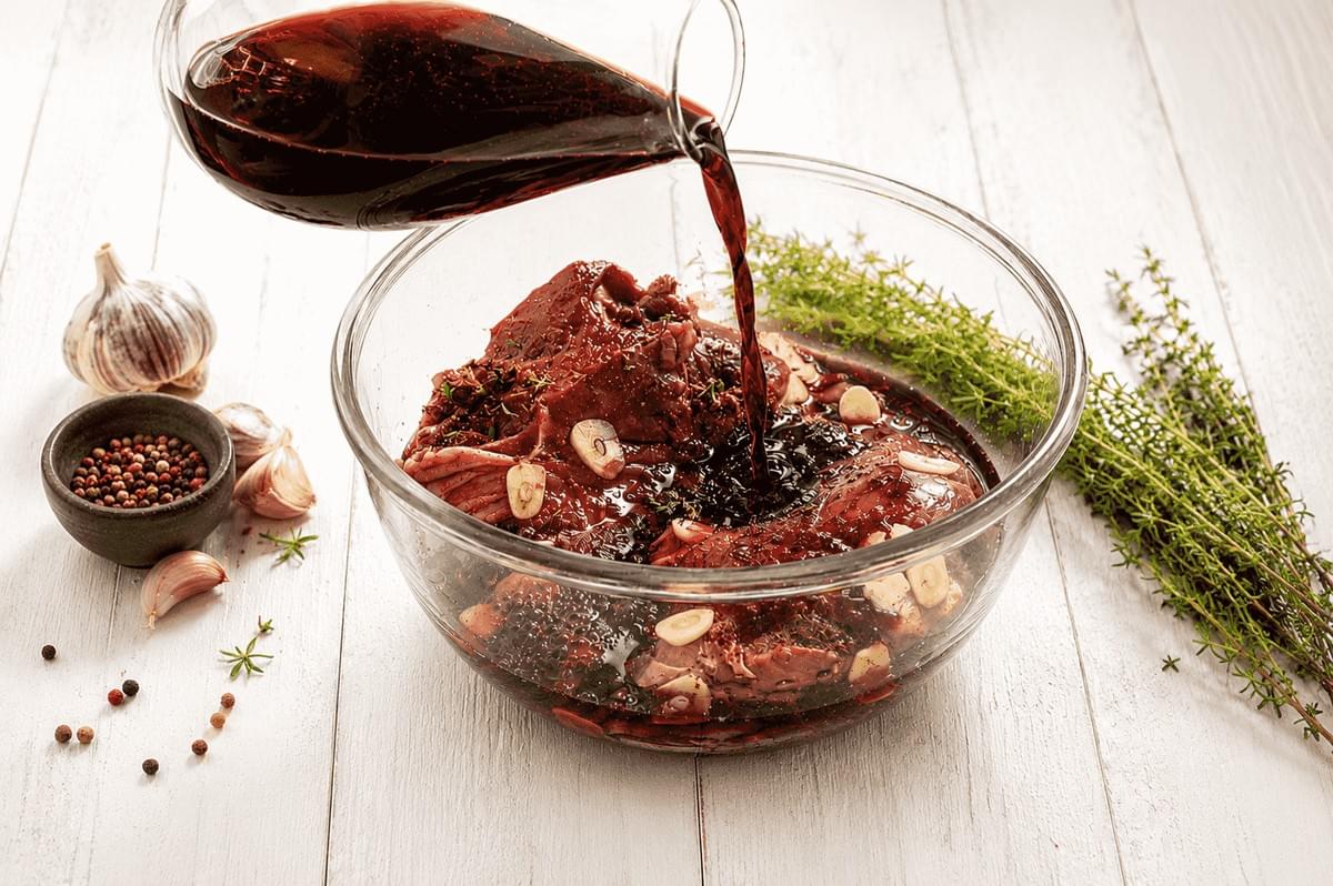 Red wine being poured from a glass decanter over pieces of raw meat marinated with sliced garlic in a clear glass bowl, set on a light wooden surface next to fresh thyme sprigs, a bulb of garlic, and a small bowl of peppercorns. Red wine being poured from a glass decanter over pieces of raw meat marinated with sliced garlic in a clear glass bowl, set on a light wooden surface next to fresh thyme sprigs, a bulb of garlic, and a small bowl of peppercorns.