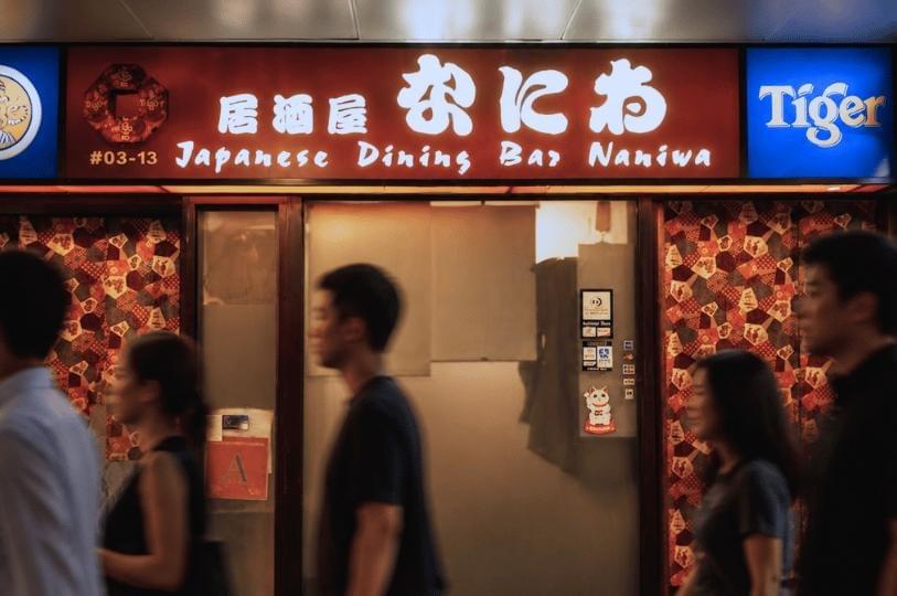 The image titled shows the brightly lit storefront of "Japanese Dining Bar Naniwa," featuring a prominent red sign with white Japanese and English lettering alongside a blue Tiger Beer logo. In the foreground, several blurred figures of people walk past the restaurant's entrance, creating a sense of movement in front of the patterned curtains and glass door. The image titled shows the brightly lit storefront of "Japanese Dining Bar Naniwa," featuring a prominent red sign with white Japanese and English lettering alongside a blue Tiger Beer logo. In the foreground, several blurred figures of people walk past the restaurant's entrance, creating a sense of movement in front of the patterned curtains and glass door.