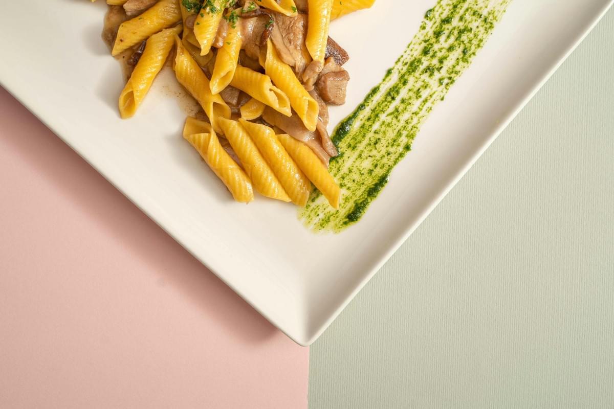 A top-down view shows a white rectangular plate featuring a serving of yellow penne pasta tossed with mushrooms and a light sauce. A vibrant streak of green herb oil or pesto runs diagonally across the plate, set against a split background of pastel pink and light grey. A top-down view shows a white rectangular plate featuring a serving of yellow penne pasta tossed with mushrooms and a light sauce. A vibrant streak of green herb oil or pesto runs diagonally across the plate, set against a split background of pastel pink and light grey.