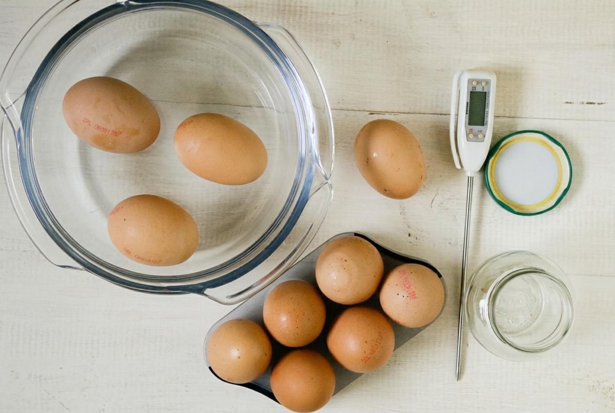 Brown eggs in a glass bowl of water and an egg carton next to a kitchen thermometer. Brown eggs in a glass bowl of water and an egg carton next to a kitchen thermometer.