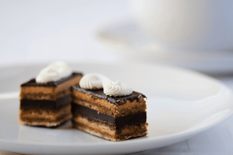 Two bite-sized, layered pieces of opera cake served on a bright white plate, each elegantly garnished with a delicate swirl of white cream. Two bite-sized, layered pieces of opera cake served on a bright white plate, each elegantly garnished with a delicate swirl of white cream.