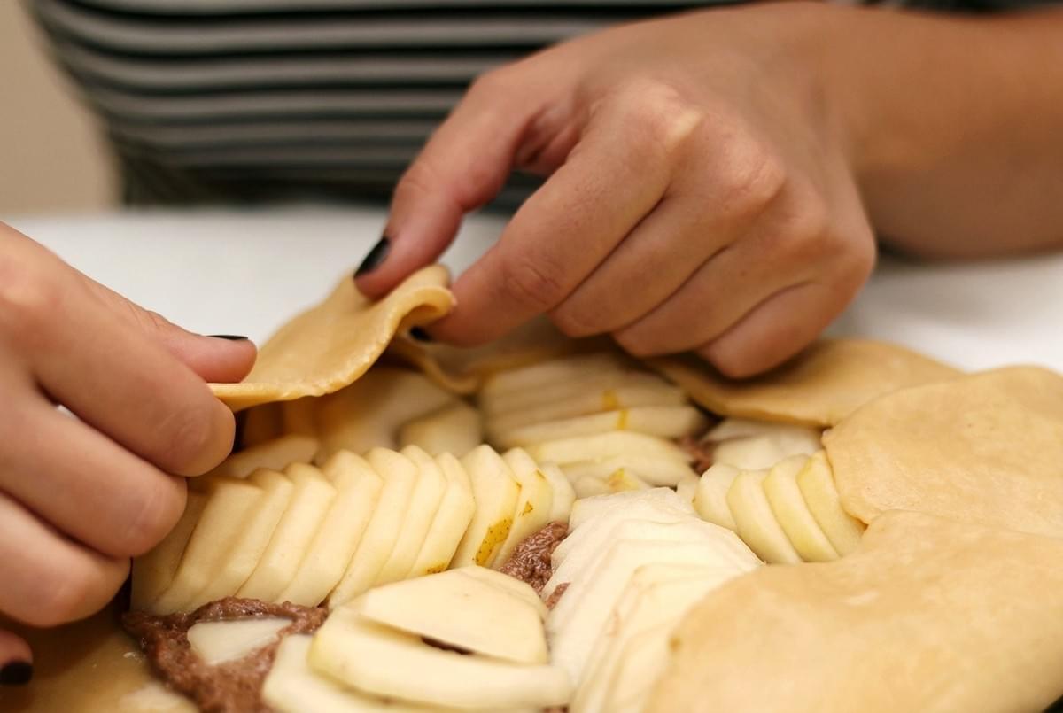 Close-up of hands folding pastry crust edges over sliced pears to form a rustically shaped galette. Close-up of hands folding pastry crust edges over sliced pears to form a rustically shaped galette.