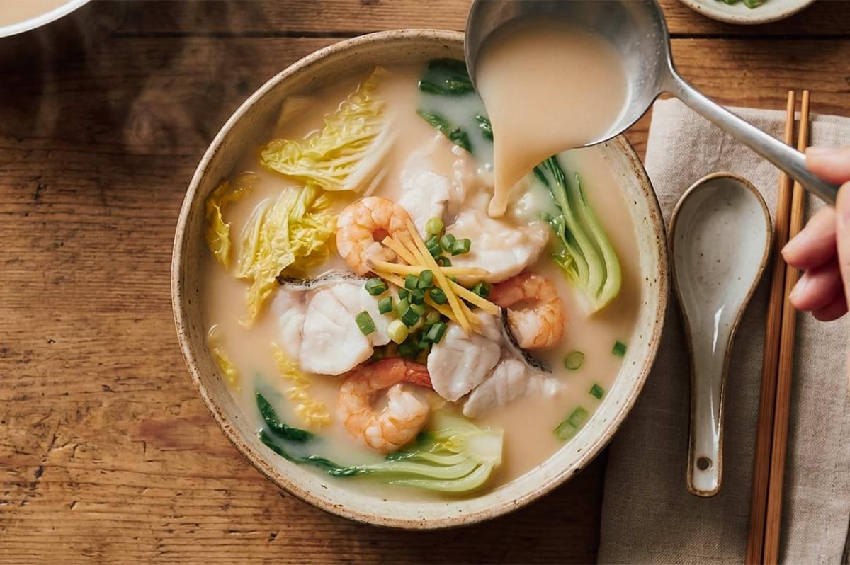 Ladle pouring rich, milky fish broth into a bowl filled with fish slices, whole prawns, napa cabbage, and ginger matchsticks. Ladle pouring rich, milky fish broth into a bowl filled with fish slices, whole prawns, napa cabbage, and ginger matchsticks.