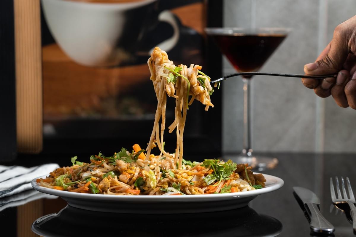 A hand uses a fork to lift a generous portion of steaming stir-fried noodles from a white plate filled with vegetables and chicken. In the blurred background, a dark cocktail in a martini glass sits on the sleek, reflective table alongside a picture of a coffee cup. A hand uses a fork to lift a generous portion of steaming stir-fried noodles from a white plate filled with vegetables and chicken. In the blurred background, a dark cocktail in a martini glass sits on the sleek, reflective table alongside a picture of a coffee cup.