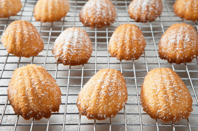 Freshly baked madeleines resting on a wire cooling rack, generously dusted with a layer of powdered sugar. Freshly baked madeleines resting on a wire cooling rack, generously dusted with a layer of powdered sugar.