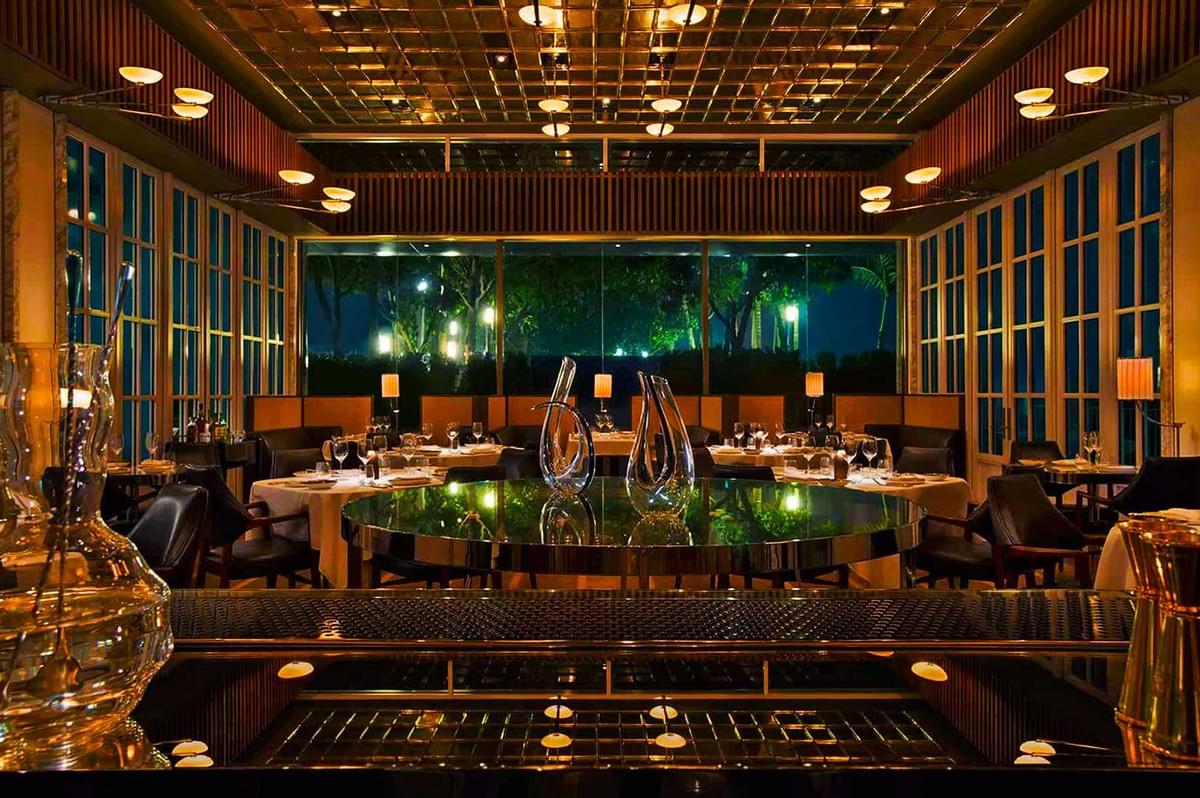 Elegant restaurant interior with dim lighting, featuring a glossy round table, sculptural vases, and candlelit tables against large windows showing greenery. Elegant restaurant interior with dim lighting, featuring a glossy round table, sculptural vases, and candlelit tables against large windows showing greenery.