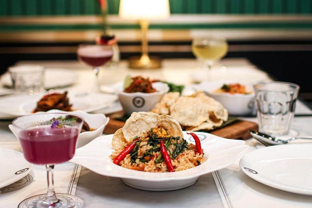 A white bowl of seasoned rice garnished with red chilies, green vegetables, and crispy crackers sits prominently on a dining table alongside a glass of purple cocktail. In the softly blurred background, additional dishes and colorful drinks are spread across the white tabletop in a restaurant setting. A white bowl of seasoned rice garnished with red chilies, green vegetables, and crispy crackers sits prominently on a dining table alongside a glass of purple cocktail. In the softly blurred background, additional dishes and colorful drinks are spread across the white tabletop in a restaurant setting.