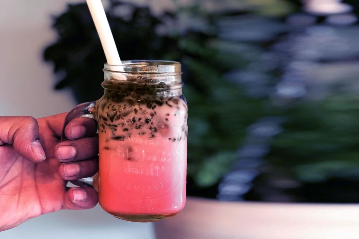 Hand holding a mason jar filled with a pink and brown layered drink, topped with seeds and a straw. Blurred green background adds a fresh feel. Hand holding a mason jar filled with a pink and brown layered drink, topped with seeds and a straw. Blurred green background adds a fresh feel.