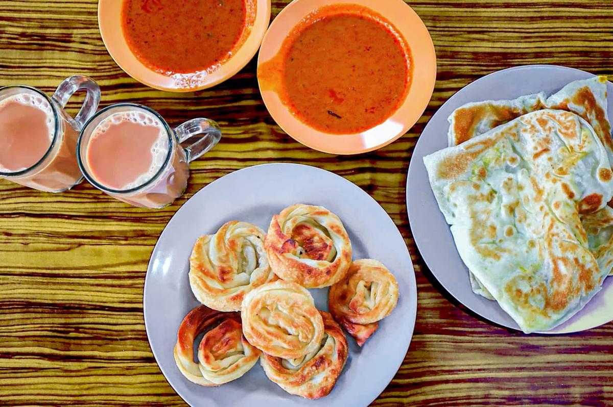A vibrant table setting features two orange bowls of rich curry, a plate of swirled pastries, folded flatbreads, and two mugs of frothy tea, conveying warmth. A vibrant table setting features two orange bowls of rich curry, a plate of swirled pastries, folded flatbreads, and two mugs of frothy tea, conveying warmth.